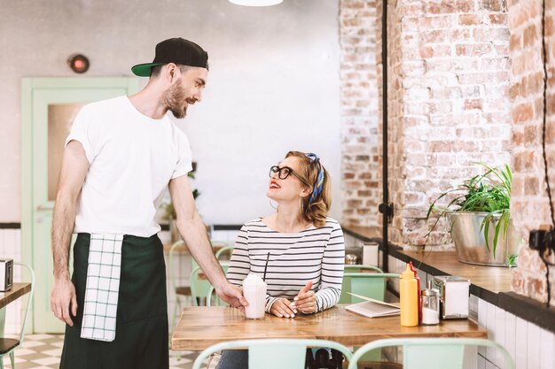 Waiter in black cap giving milkshake to pretty smiling lady in eyeglasses which happily looking at him while sitting at the table in cafe