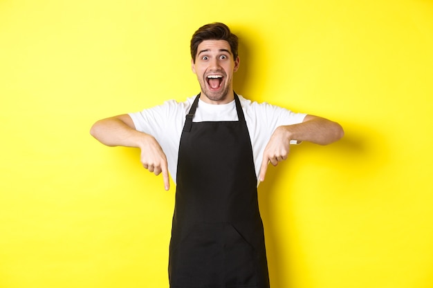 Free photo waiter, coffee shop barista in black apron pointing fingers down, looking amazed, standing over yellow background.