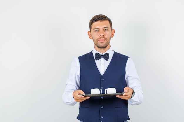 Free photo waiter holding cups in tray in shirt, vest, bow tie and looking serious. front view.