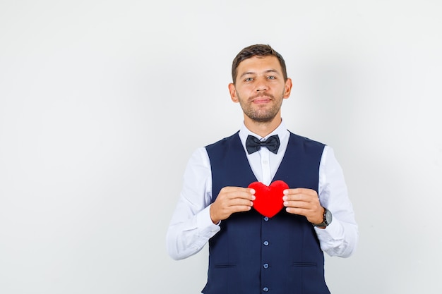 Waiter holding red heart in shirt, vest and looking cheerful. front view.