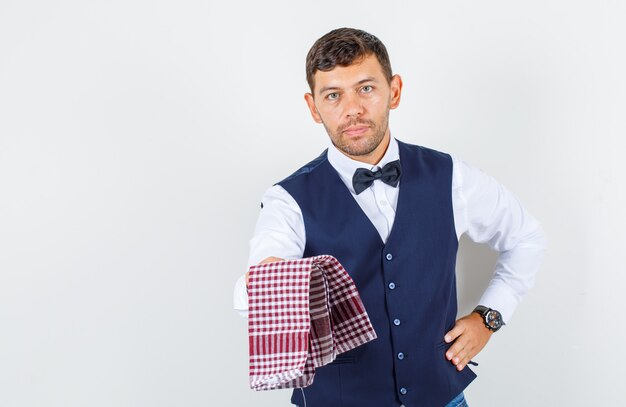 Waiter offering checked towel in shirt, vest, jeans and looking serious , front view.