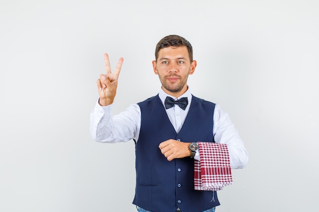 Waiter in shirt, vest, bow tie showing victory sign and looking glad , front view.