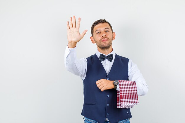 Waiter showing palm up in shirt, vest, jeans and looking confident , front view.