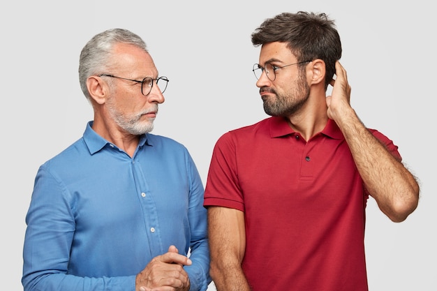 War of generations concept. Displeased bearded mature father and son look angrily at each other, have argue, cant find common solution, pose against white wall. Bad family relations.