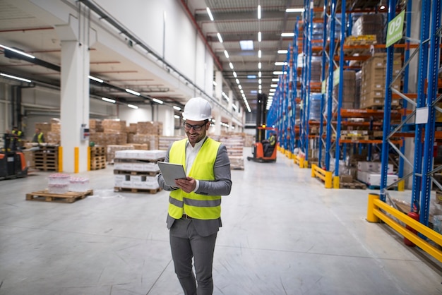 Free photo warehouse manager walking through large storage area and holding tablet while forklift operating in background