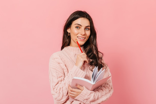 Wavy-haired lady with snow-white smile looks into camera. Girl in knitted sweater with notebook in her hands posing on isolated background.