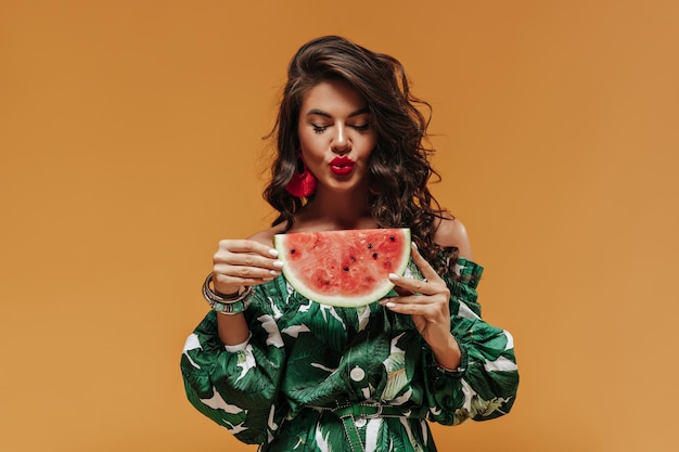 Wavy long haired trendy girl with bright lipstick and unusual accessories in green summer clothes posing with watermelon