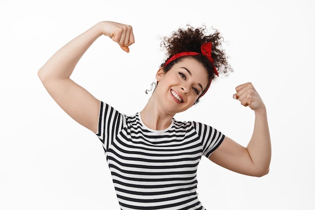 Free photo we can do it women rights strong and confident brunette girl flex biceps smiling and showing strong hands arm muscles standing against white background