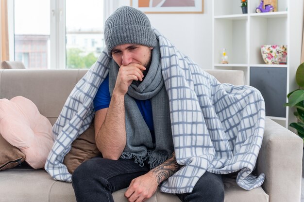 Weak and aching young ill man wearing scarf and winter hat wrapped in blanket sitting on sofa in living room looking down keeping hand on mouth