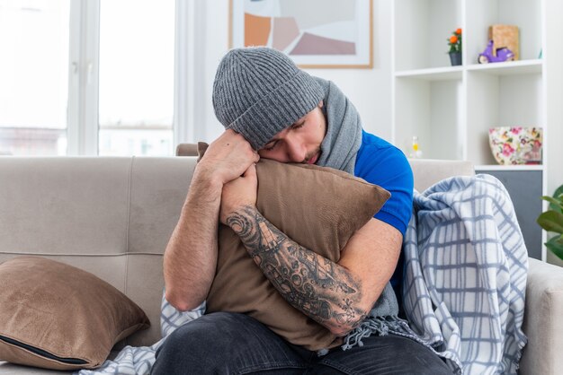 weak and tired young ill man wearing scarf and winter hat sitting on sofa in living room hugging pillow resting head on it with closed eyes