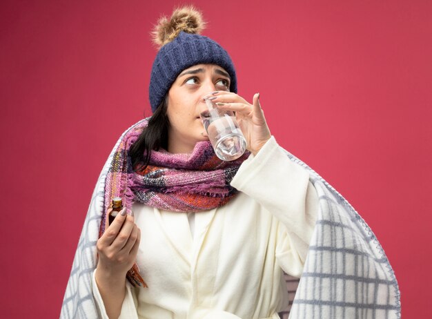 Weak young caucasian ill woman wearing robe, winter hat and scarf, drinking glass of water mixed with medicament