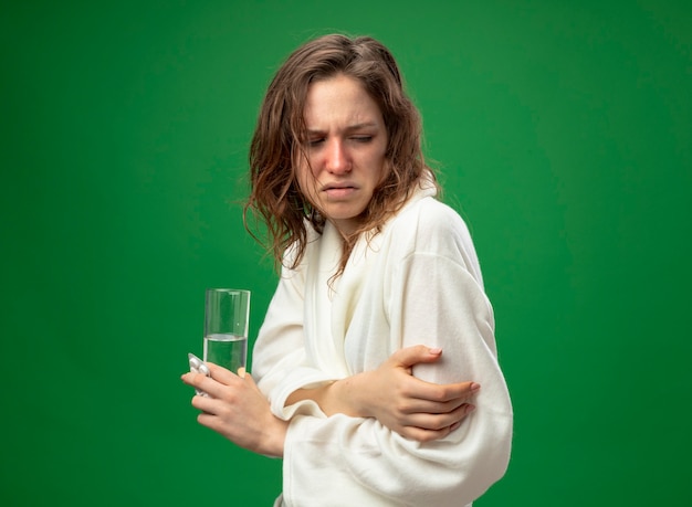 Free photo weak young ill girl looking down wearing white robe holding glass of water crossing hands isolated on green