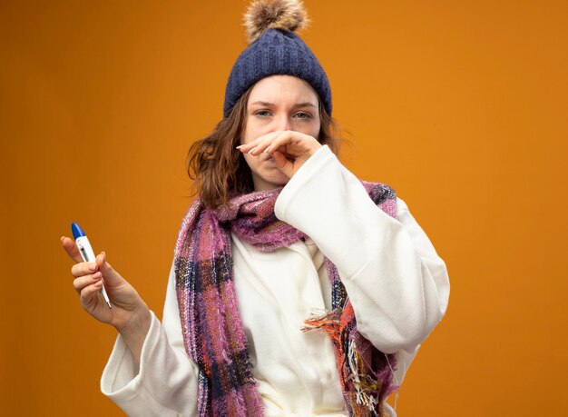 Weak young ill girl wearing white robe and winter hat with scarf holding thermometer wiping nose with hand isolated on orange