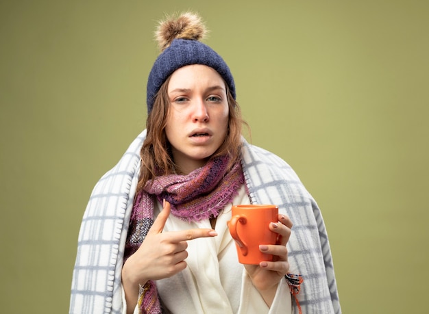 Free Photo weak young ill girl wearing white robe and winter hat with scarf wrapped in plaid holding and points at cup of tea isolated on olive green