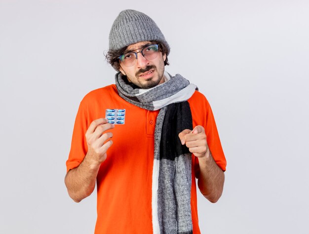 Weak young ill man wearing glasses winter hat and scarf holding pack of medical capsules looking and pointing at front isolated on white wall