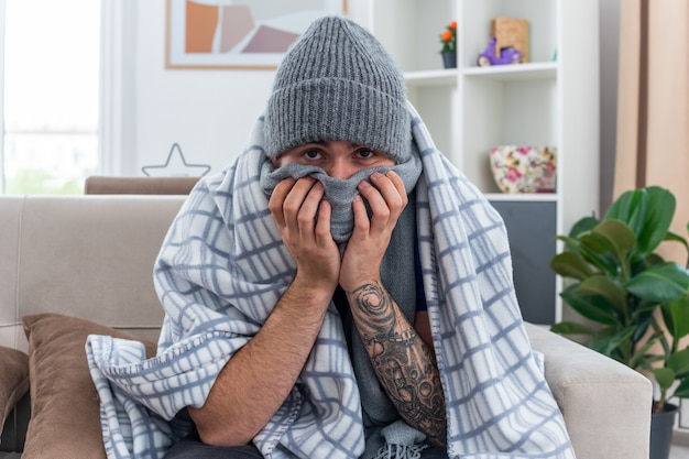 Weak young ill man wearing scarf and winter hat sitting on sofa in living room wrapped in blanket looking at camera covering mouth with scarf