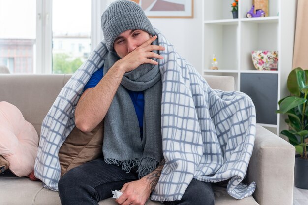 Weak young ill man wearing scarf and winter hat wrapped in blanket sitting on sofa in living room holding napkin looking at camera wiping nose with hand with one eye closed