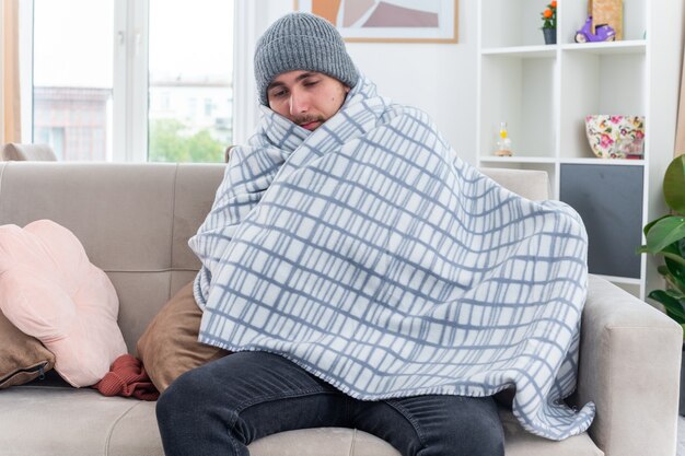 weak young ill man wearing scarf and winter hat wrapped in blanket sitting on sofa in living room looking down