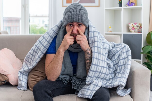 weak young ill man wearing scarf and winter hat wrapped in blanket sitting on sofa in living room looking at front pointing fingers on nose