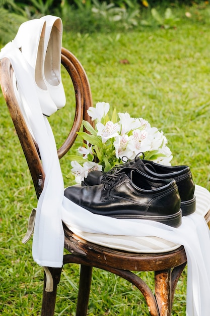 Free photo wedding black shoes and white high heels with flower bouquet on wooden chair in the garden