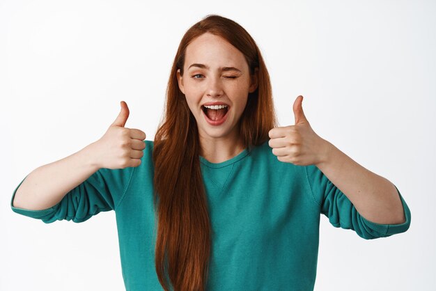 Well done Smiling redhead girl winking and show thumbs up like and approve praise nice job great work gesture hinting on something good recommend service