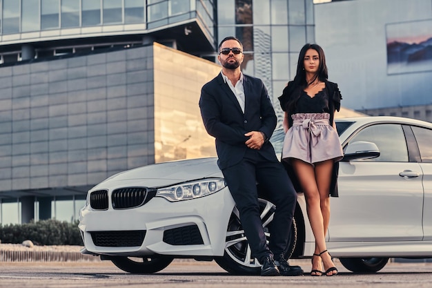 Free photo well-dressed attractive couple leaning on a luxury car outdoors against the skyscraper.