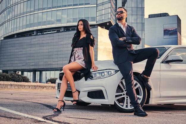 Well-dressed attractive couple leaning on a luxury car outdoors against the skyscraper.