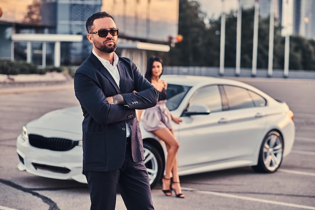 Free photo well-dressed attractive couple leaning on a luxury car outdoors against the skyscraper.