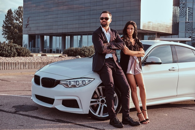 Well-dressed attractive couple leaning on a luxury car outdoors against the skyscraper.