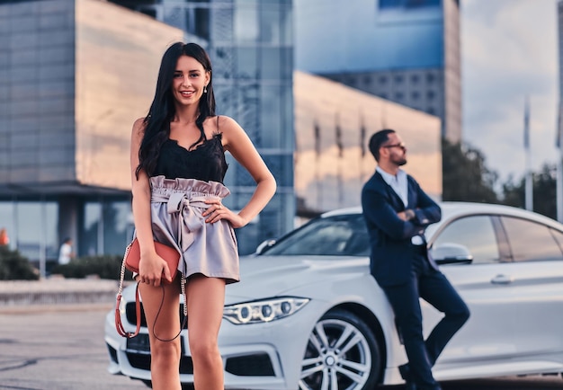 Well-dressed beautiful woman and brutal male in a suit near a luxury car outdoors against a cityscape.