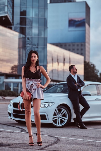 Free photo well-dressed beautiful woman and brutal male in a suit near a luxury car outdoors against a cityscape.