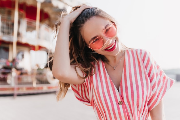 Well-dressed cute girl touching her hair while posing near carousel. White blissful woman in sunglasses spending weekend in summer amusement park.