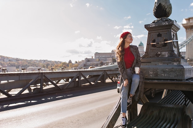 Free photo well-dressed female tourist drinking coffee on city background in windy day