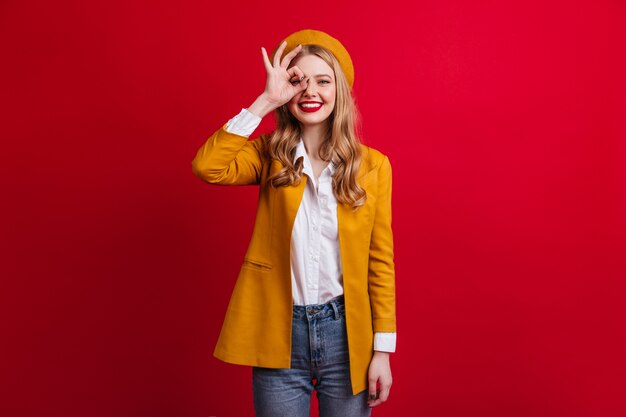 Well-dressed girl in beret showing okay sign.  female model in yellow jacket isolated on red wall.