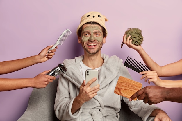 Free Photo well groomed positive man wears bathrobe, hat, uses smartphone after taking shower