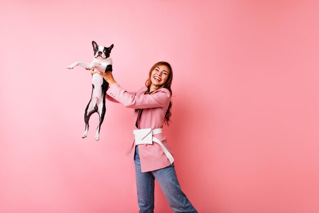 Welldressed young woman holding french bulldog and laughing to camera Studio photo of beautiful white girl playing with dog on pastel background