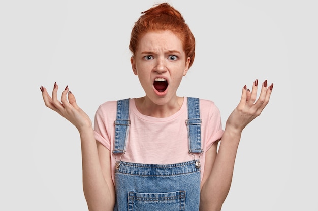 Free photo what hell! depressed puzzled red haired woman with angry annoyed facial expression, spreads hands, exclaims with exasperation, has enraged look, isolated over white wall. negative emotions