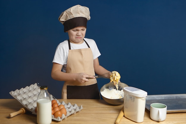 What a mess. Disappointed male kid trying to beat eggs with flour and milk while making dough by himself for the first time but having some problem