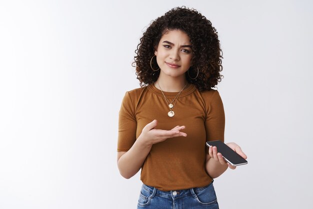 What was message about. Confused clueless pretty modern 20s curly dark hair girl hold smartphone pointing phone shrugging clueless raise eyebrow curious puzzled receive strange text white background