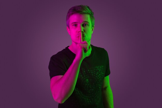 Whispering a secret. Copyspace. Caucasian man's portrait on purple studio background in neon light. Beautiful male model in black shirt. Concept of human emotions, facial expression, sales, ad.