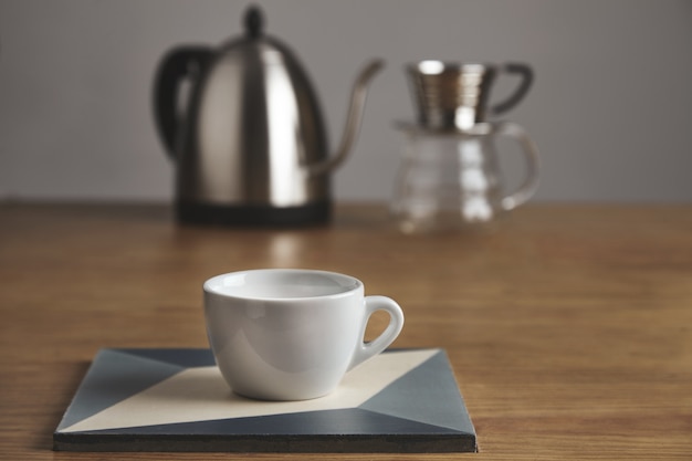 White blank coffee cup in front of modern teapot and beautiful transparent drip coffee maker.
Cup on ceramic plate on thick wooden table in cafe shop.