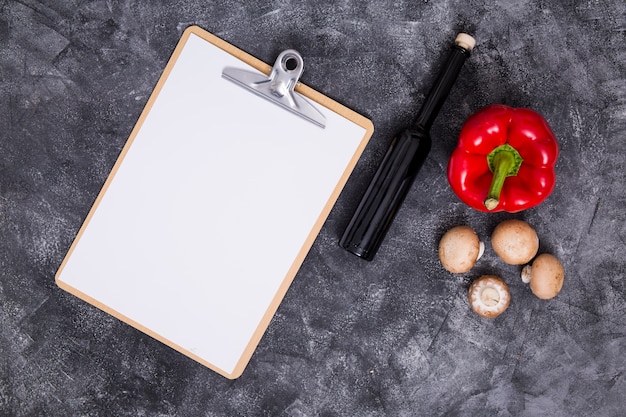 White blank paper on clipboard with bell pepper; mushroom and bottle on black textured backdrop