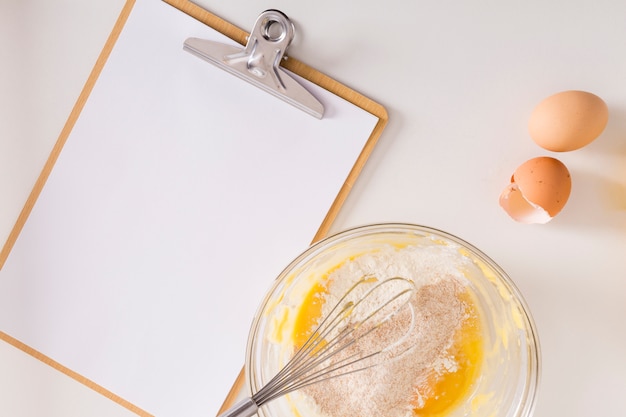 White blank paper on clipboard with whipped egg and flour bowl on white backdrop