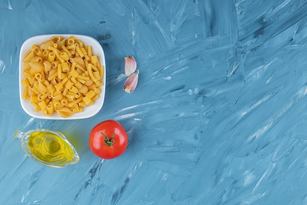 A white board of raw pasta with oil and fresh red tomatoes on a blue background. 