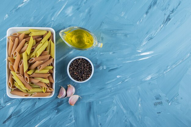 A white board of raw pasta with oil and pepper corns on a blue background. 