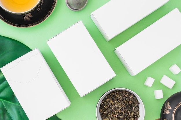 White boxes with healthy tea with herbs and sugar cubes on green backdrop