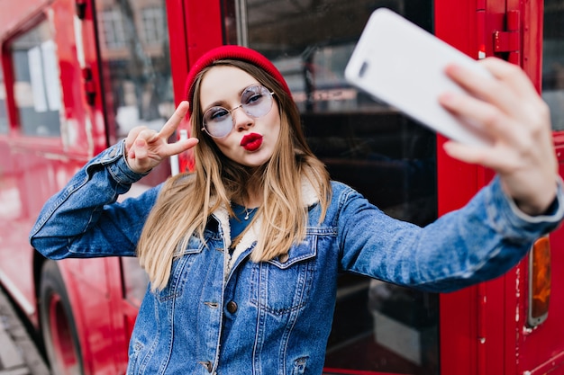 White caucasian girl using phone for selfie while making kissing face expression. Outdoor lady in denim jacket and red hat standing on the street and taking picture of herself.