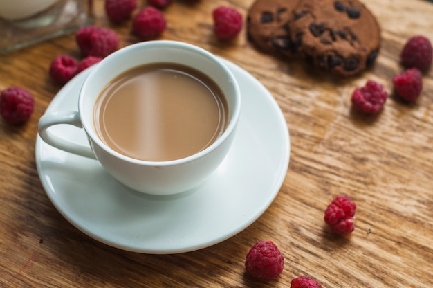 Free photo white cup of coffee with chocolate cookies and raspberries on wooden background