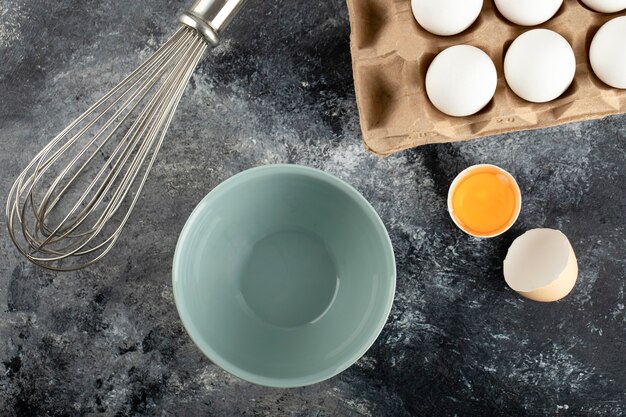 White eggs in carton container, bowl and whisker on marble surface. 