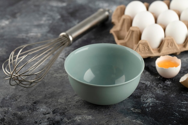 White eggs in carton container, bowl and whisker on marble surface. 